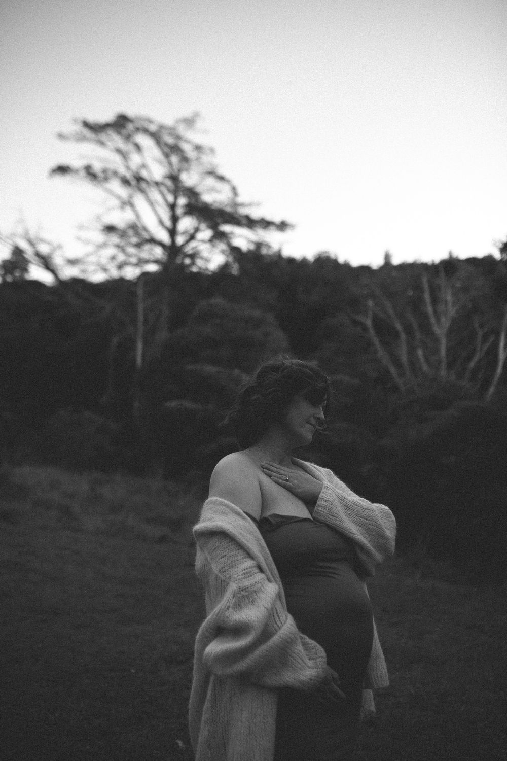 Black and white photo of a pregnant woman in a fur wrap standing outdoors at dusk with trees in the background.