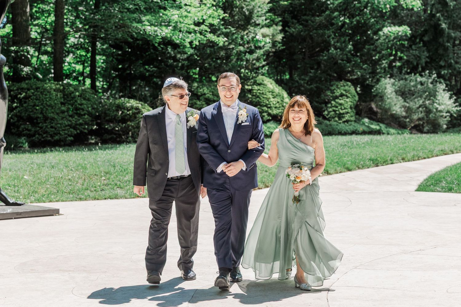 Bride in sage green dress walks with two men in suits at outdoor wedding ceremony.