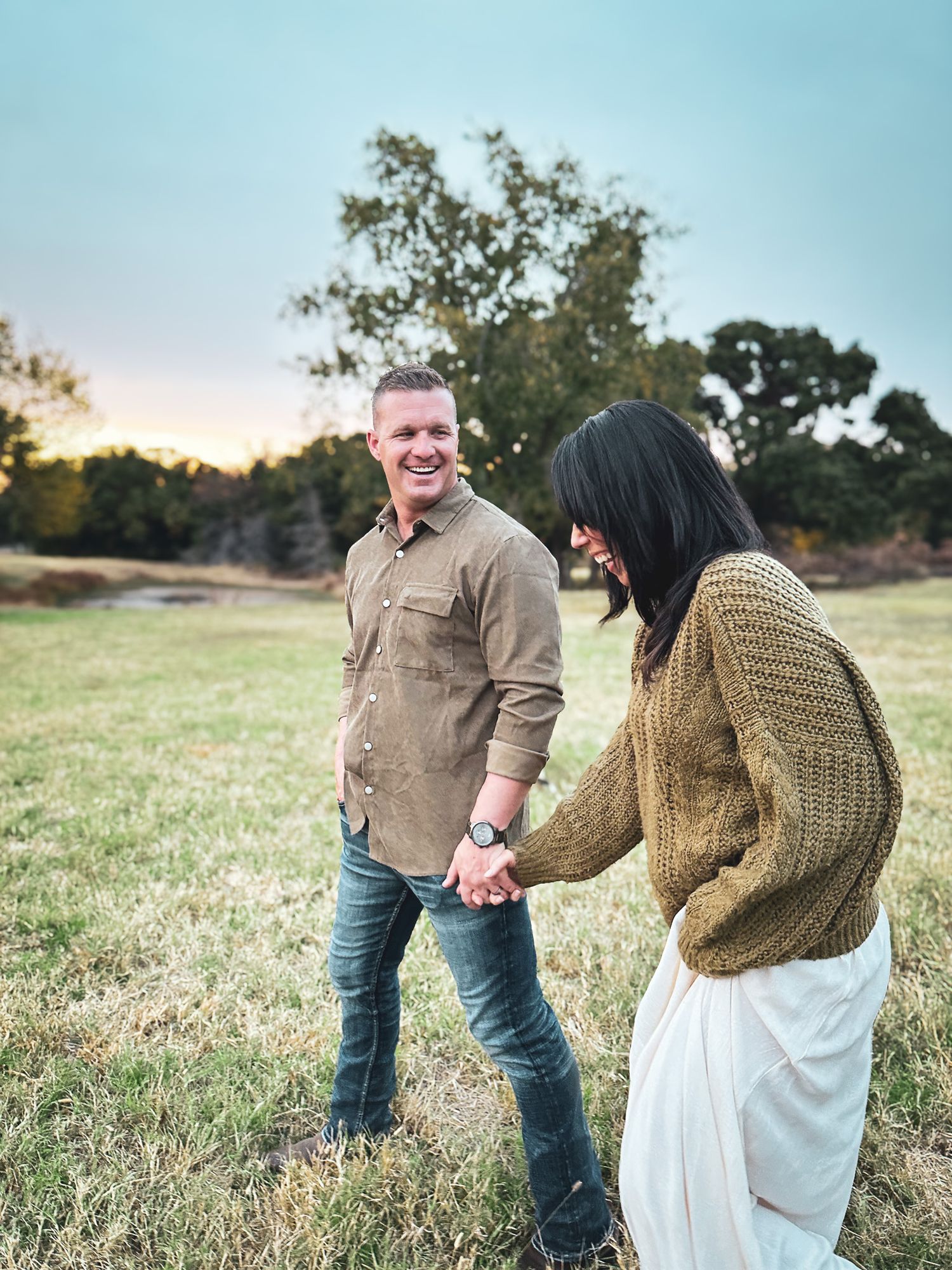 Smiling couple holding hands in a grassy field at sunset with trees in the background.