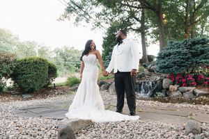 Bride in white mermaid gown and groom in white tuxedo hold hands on a garden path with waterfall backdrop.