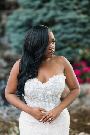 A Black bride in a strapless white floral appliqué wedding gown poses gracefully outdoors with lush greenery behind her.