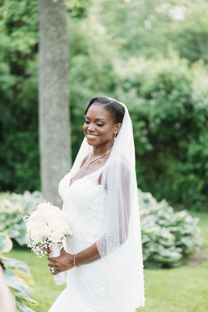 Smiling Black bride in white lace gown and veil holds white bouquet outdoors in lush garden setting.