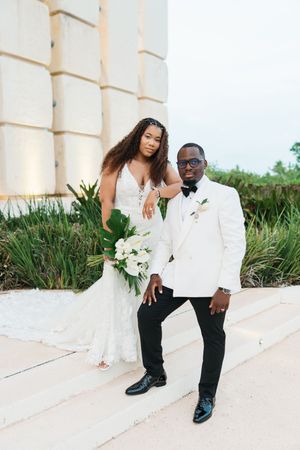 Black bride and groom pose in white wedding attire, she holds tropical bouquet, outdoors near stone building.