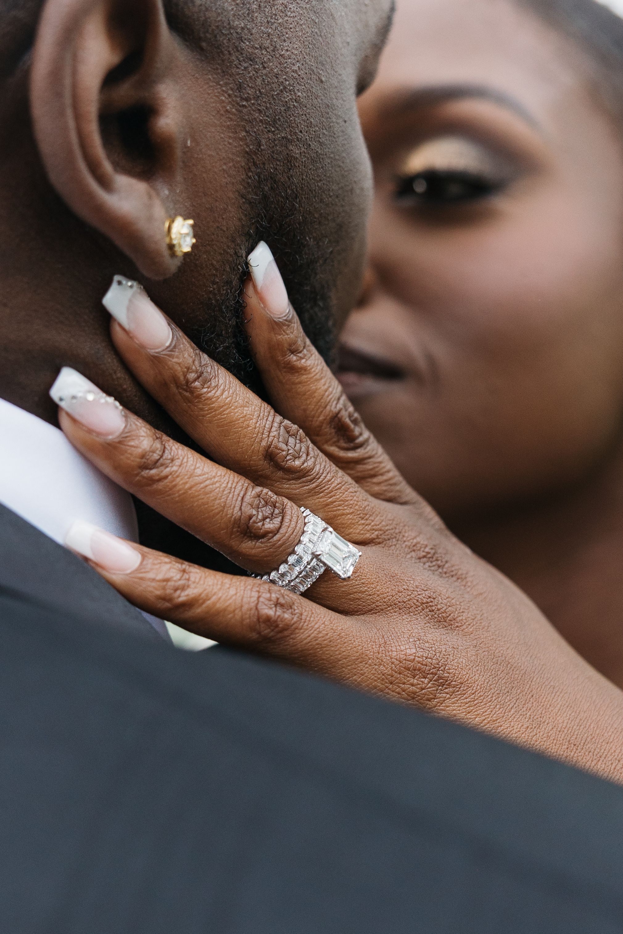 A bride with diamond rings tenderly touches her groom's face, showcasing elegant nail art during a romantic wedding moment.