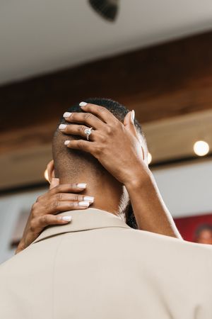 A woman with white nails and a diamond ring tenderly cradles a man's head from behind in a warm indoor setting.
