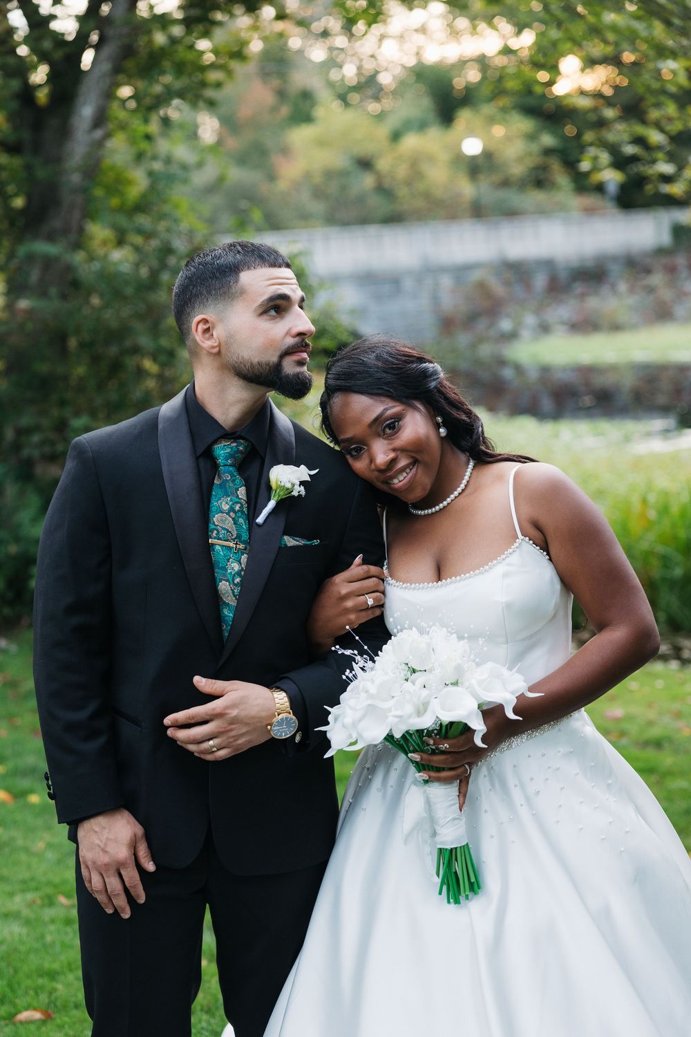 Interracial couple in wedding attire, groom in black suit with green tie, bride in white gown holding bouquet outdoors.