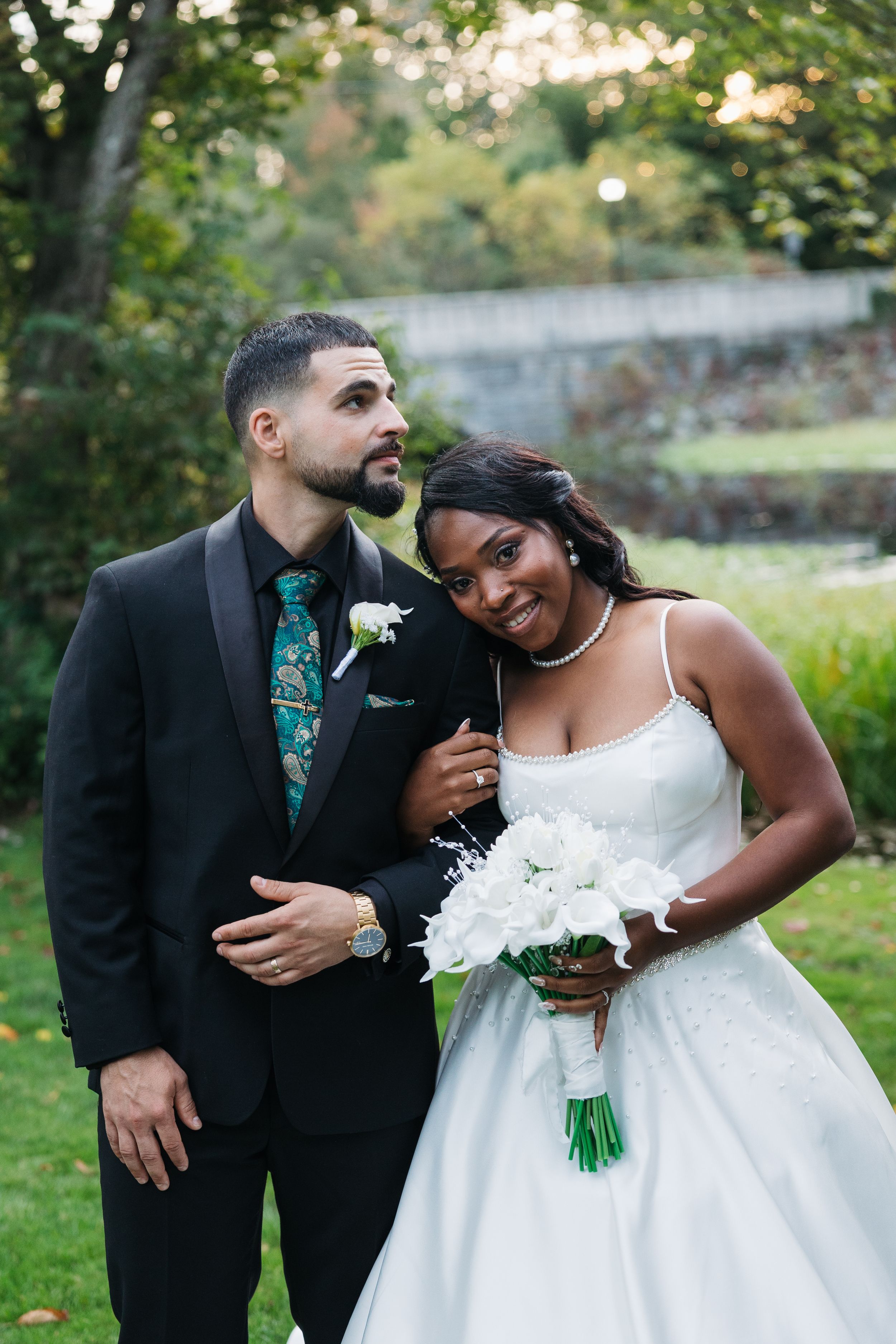 Interracial couple in wedding attire, groom in black suit with green tie, bride in white gown holding bouquet outdoors.
