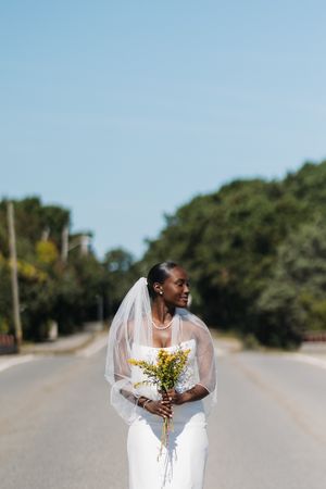 A Black bride in a white gown and veil holds yellow wildflowers while standing on a tree-lined road under a clear blue sky.