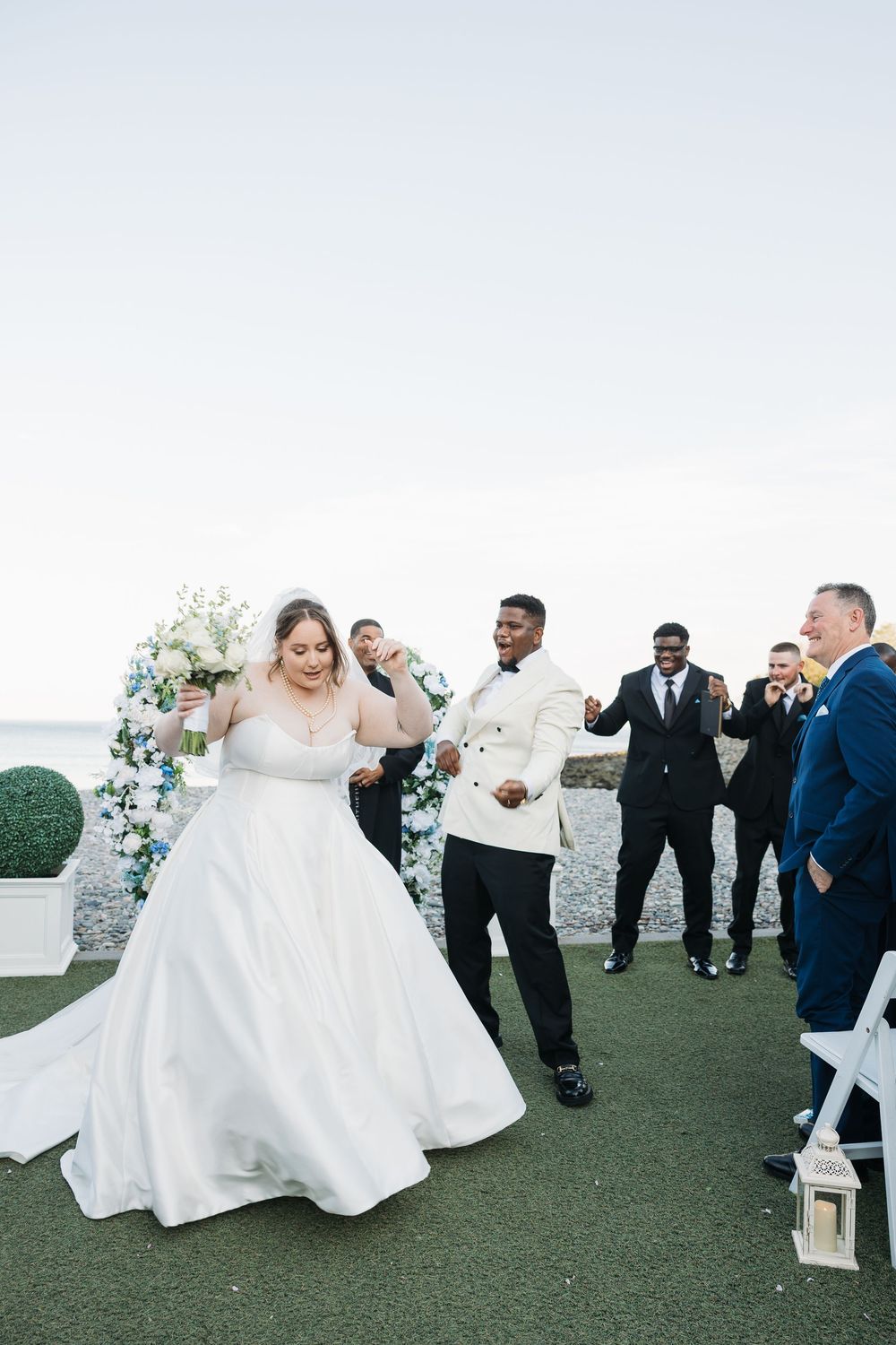 Bride in white gown and groom in white tuxedo walk down the aisle at an outdoor seaside wedding ceremony.