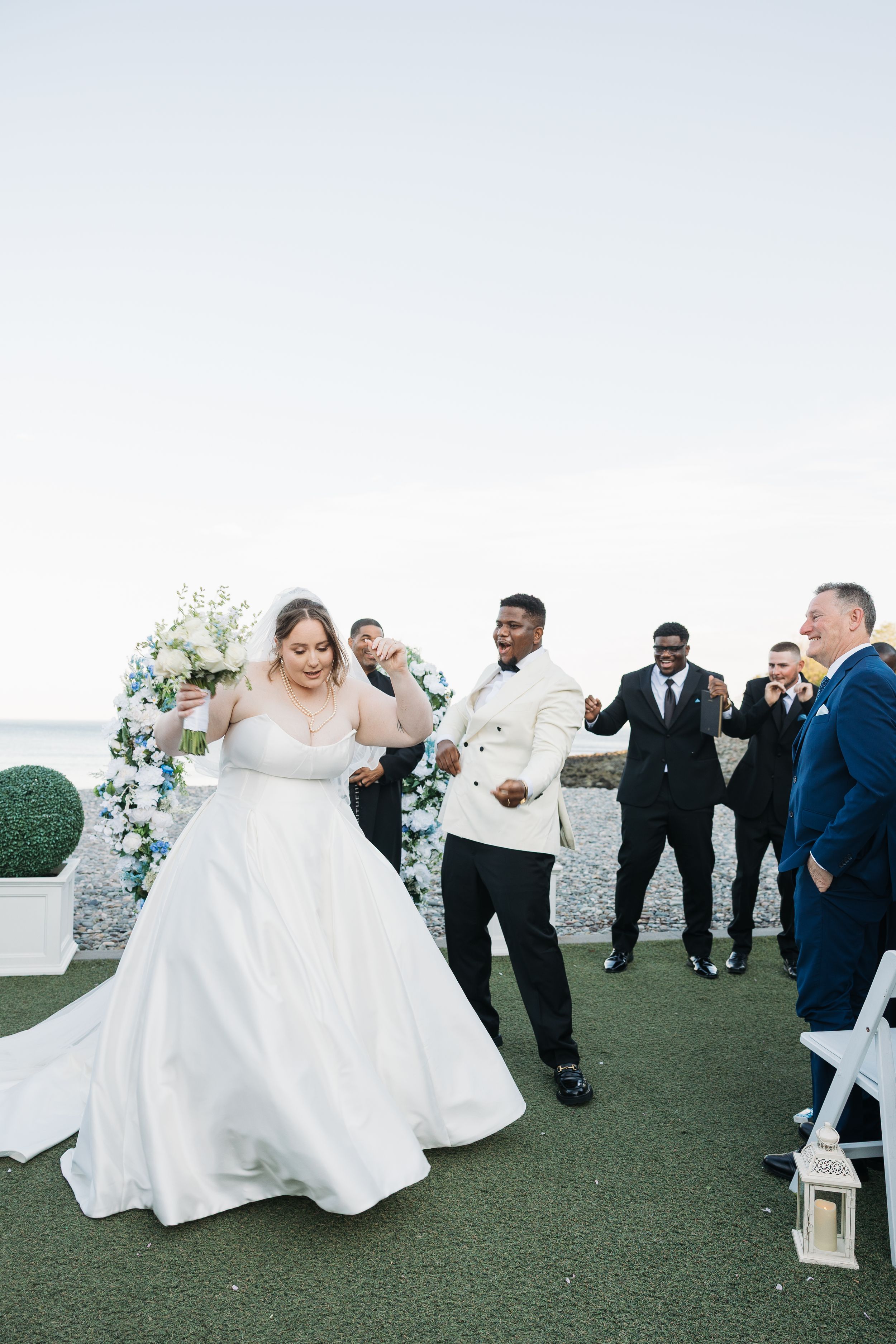 Bride in white gown and groom in white tuxedo walk down the aisle at an outdoor seaside wedding ceremony.