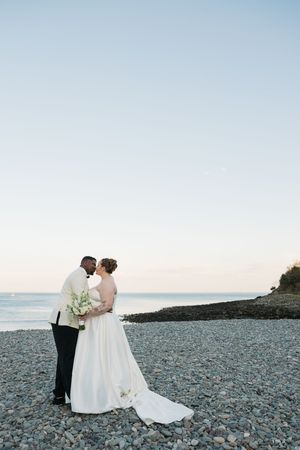 Bride and groom share a romantic moment on a rocky beach at sunset, with ocean waves in the background.