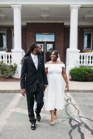 A couple in formal attire walks together outside a brick building with white columns, the woman in a white dress and man in a black suit.
