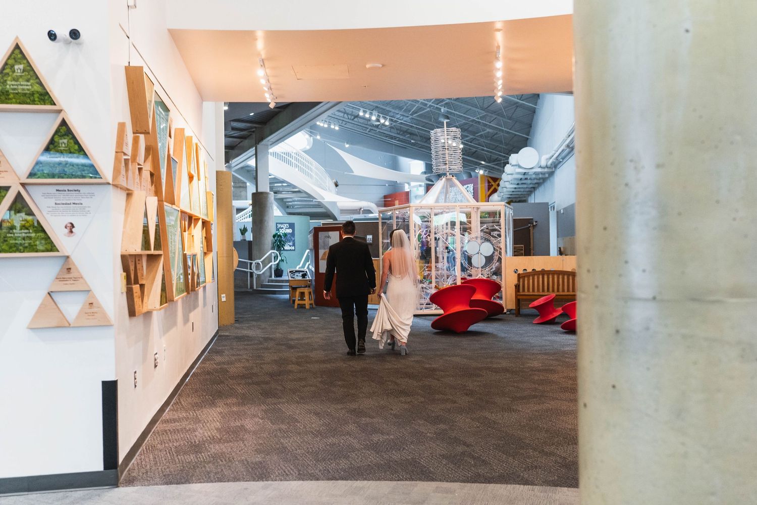 Two people walk through a modern open-plan space with art displays, red furniture, and triangular wooden shelving units.