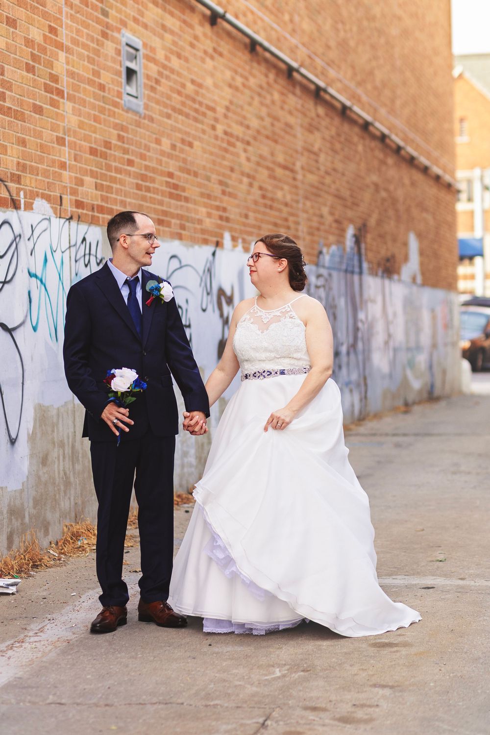 Bride and groom holding hands in front of a graffiti brick wall, smiling at each other on their wedding day.