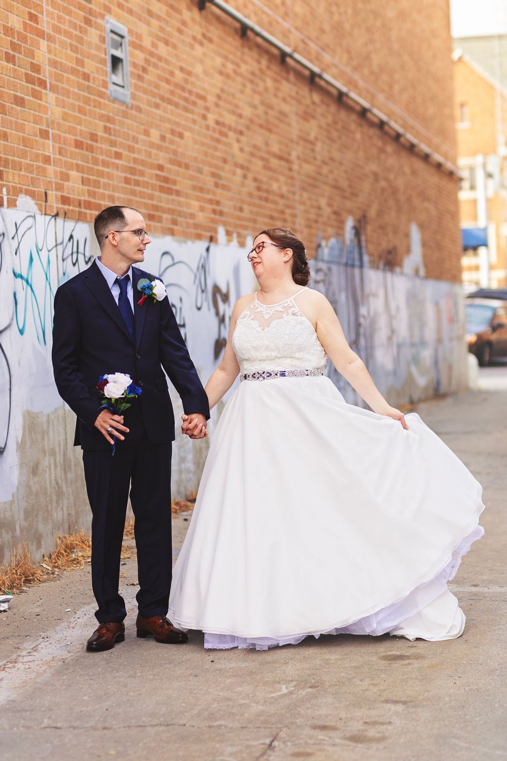 Bride in white lace gown and groom in navy suit hold hands in urban alley with brick wall and graffiti backdrop.