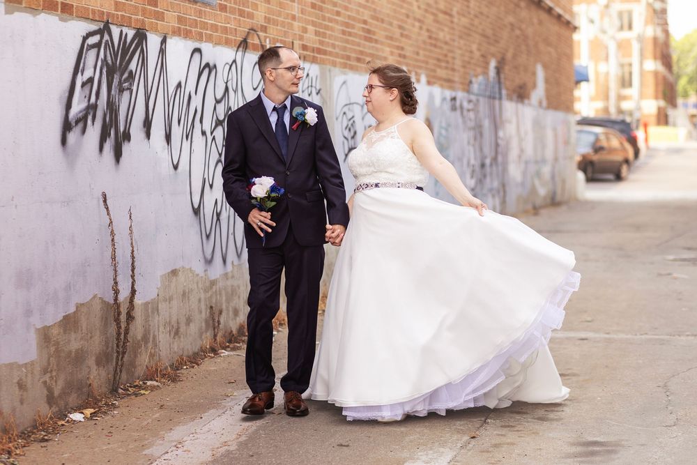 Bride in white gown twirling beside groom in navy suit, walking past graffiti wall on urban sidewalk.
