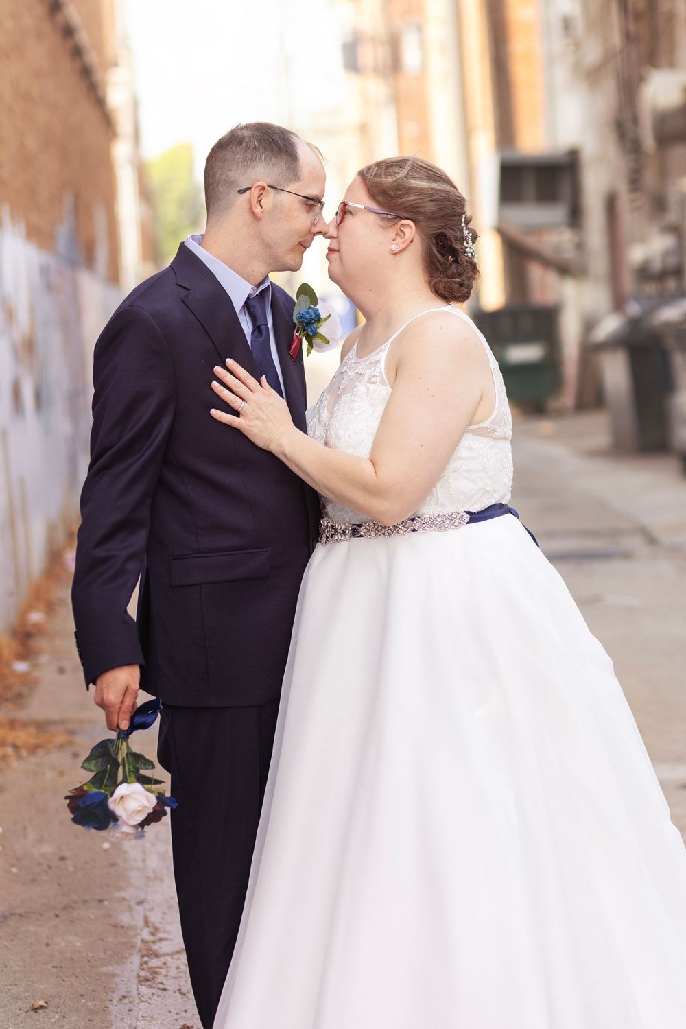 A bride and groom share a romantic moment in an urban alley, dressed in a white gown and navy suit with boutonniere.