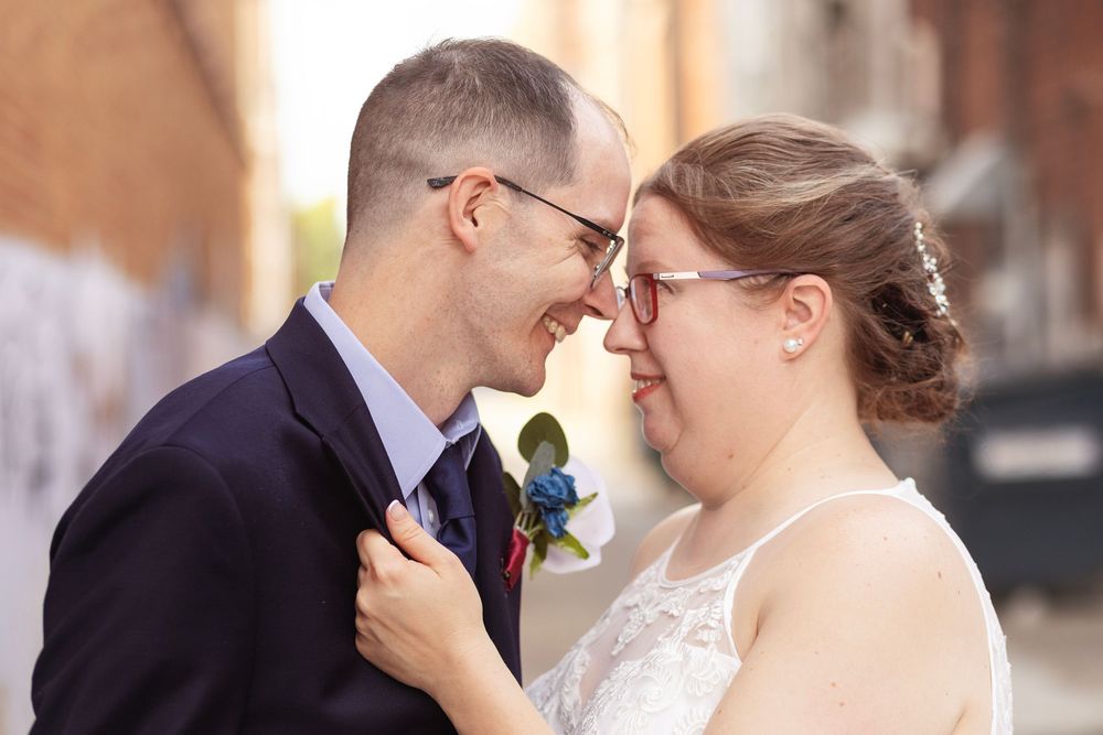 Bride and groom in glasses share a romantic nose-to-nose moment outdoors, she in lace dress, he in navy suit with boutonniere.