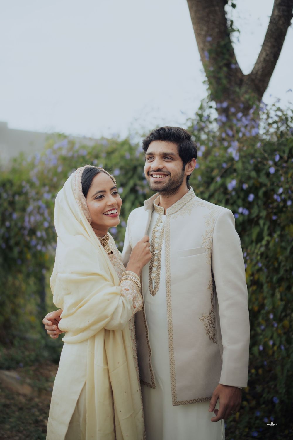 Couple in matching cream traditional Indian wedding attire posing outdoors, woman in dupatta, man in sherwani.