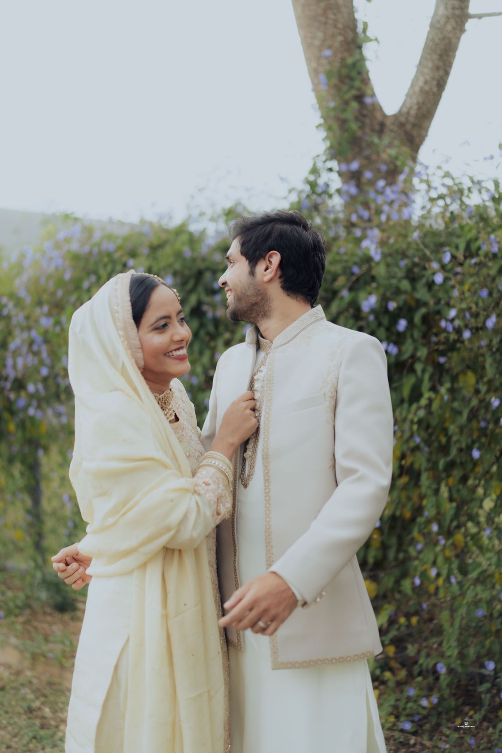 Indian couple in traditional white wedding attire smiling at each other in a lush garden setting.