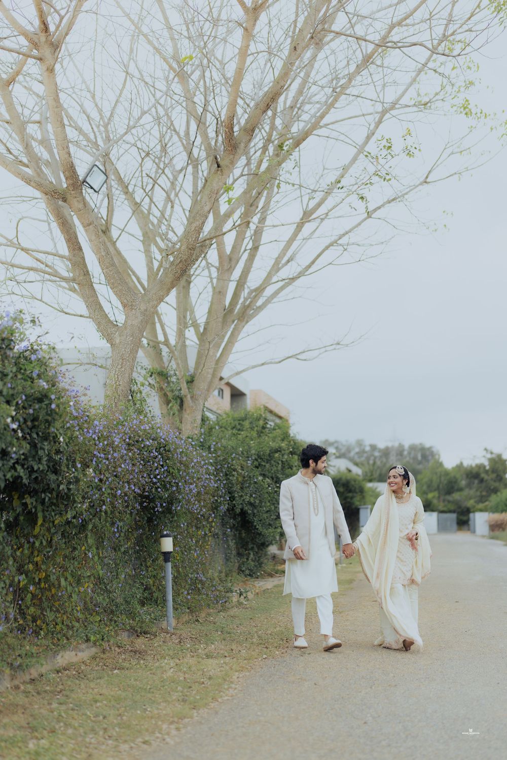 South Asian couple in white wedding attire holding hands while walking along a tree-lined path.