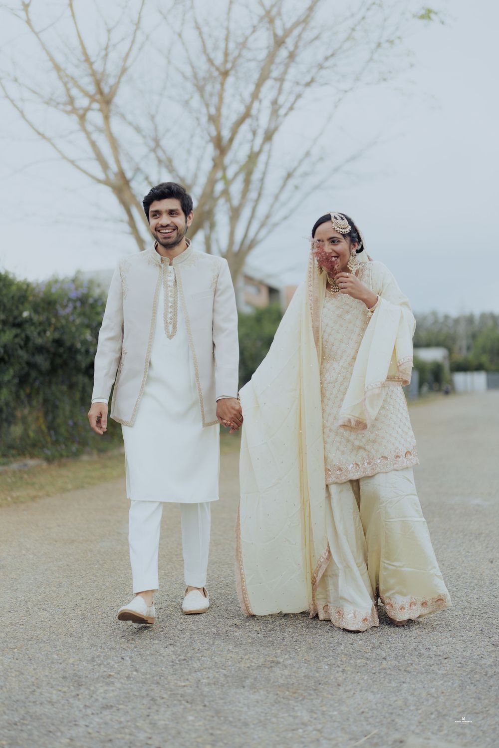 South Asian bride and groom in ivory wedding outfits holding hands and smiling on an outdoor path.