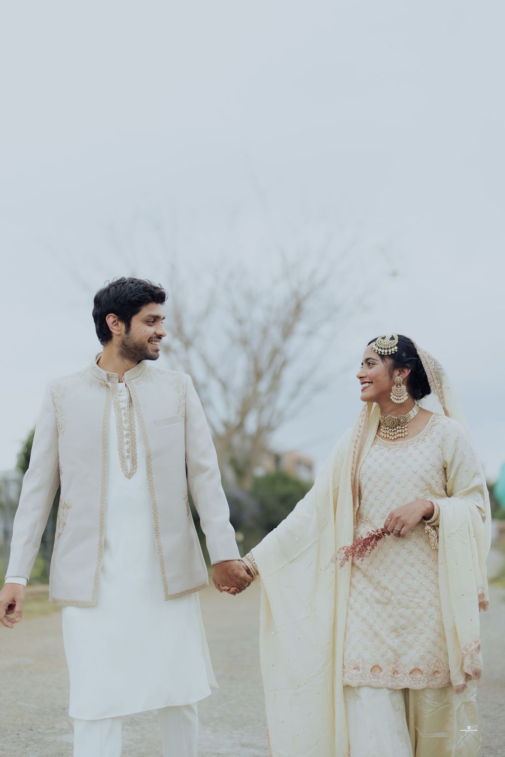 Indian couple in ivory wedding attire holding hands outdoors, bride in embroidered salwar with gold jewelry, groom in sherwani.