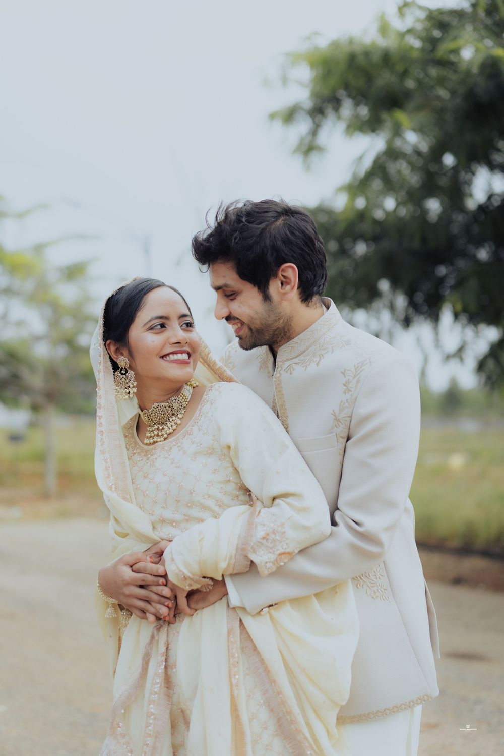 Couple in ivory Indian wedding attire embracing outdoors, woman in saree with gold jewelry, man in sherwani smiling.