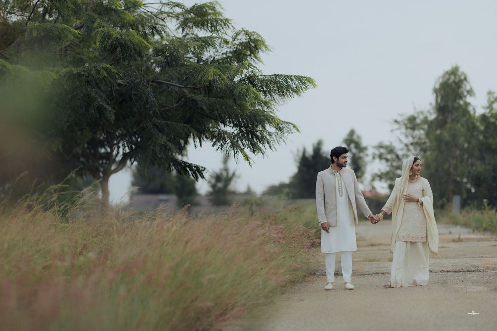 Couple in traditional South Asian attire holding hands on a dirt path surrounded by lush greenery.