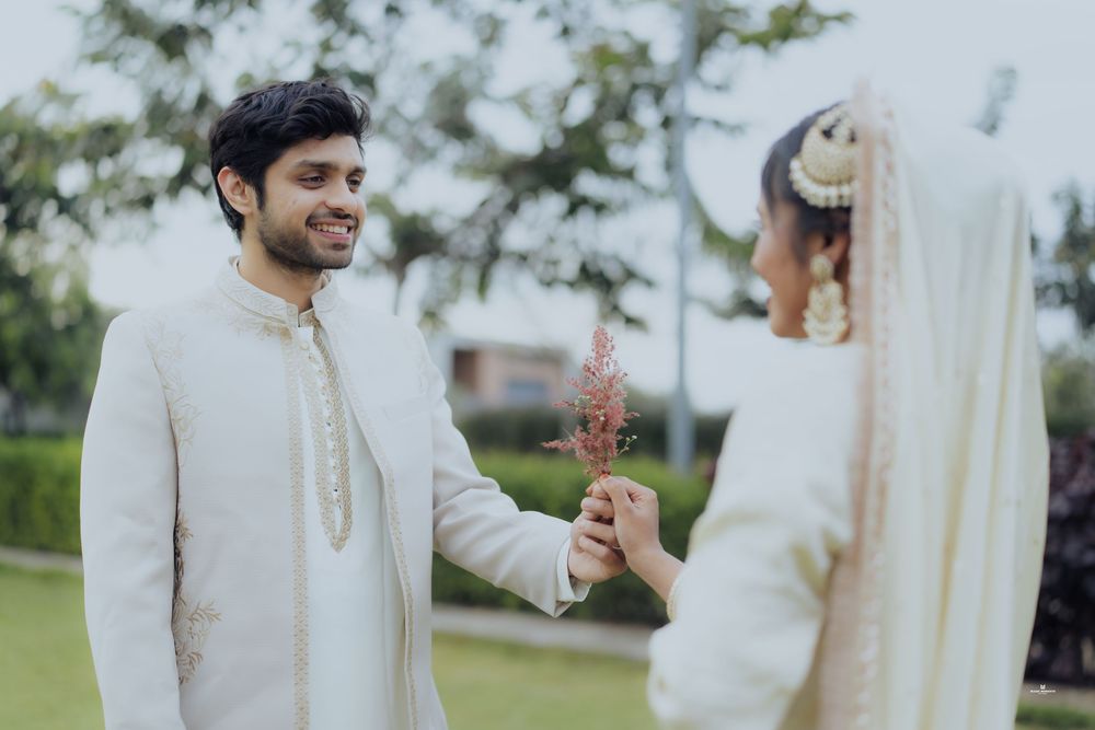 Indian groom in white sherwani smiles at bride in hijab holding pink flowers during wedding ceremony.