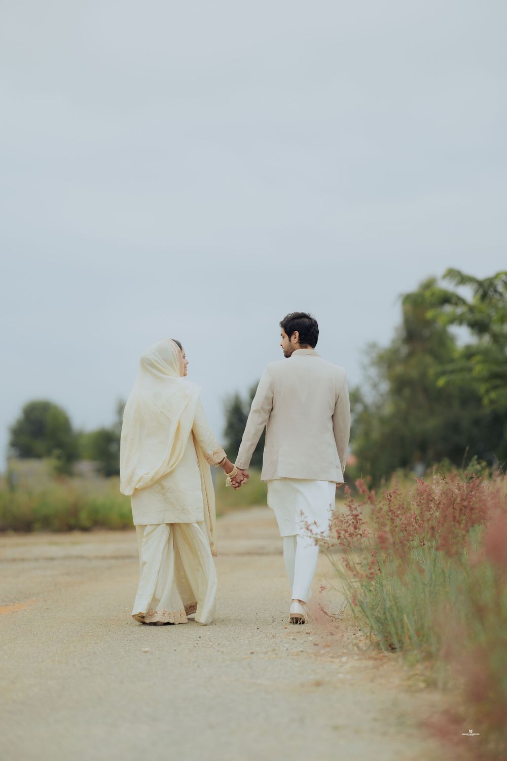 Couple in white traditional South Asian attire holding hands while walking down a scenic rural path.