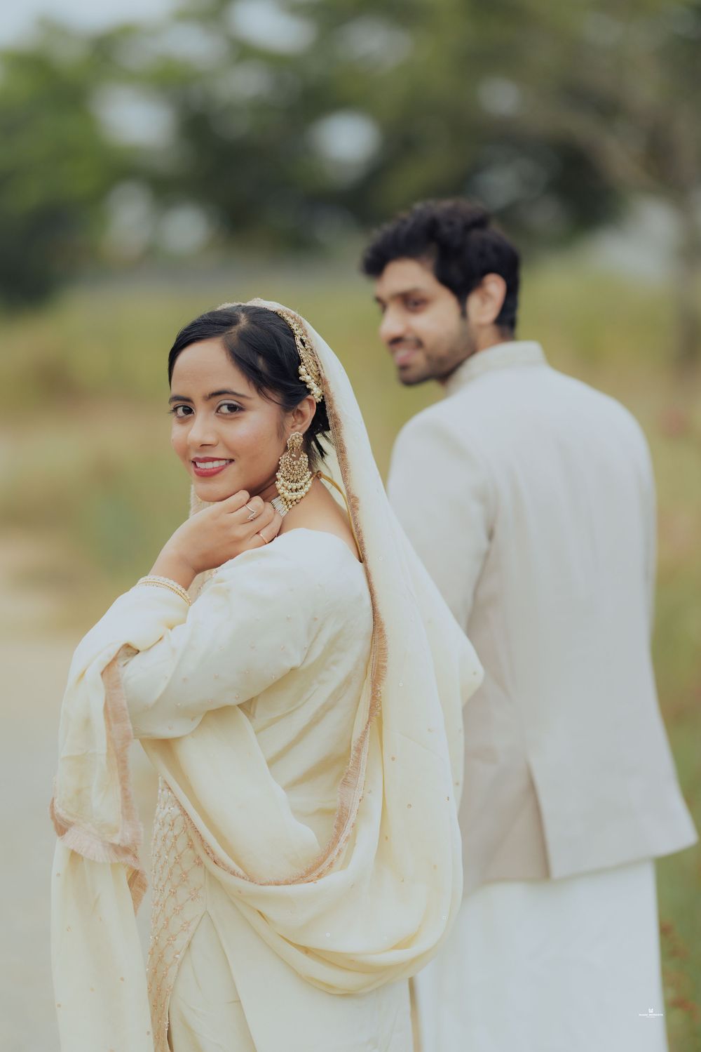 Indian couple in white traditional attire posing outdoors, bride smiling over shoulder wearing dupatta and gold jewelry.
