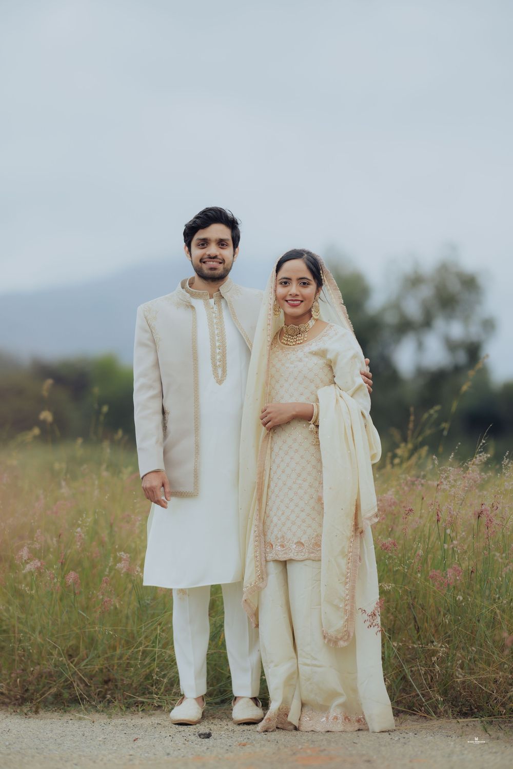 South Asian couple in matching cream traditional wedding attire posing outdoors in a field.
