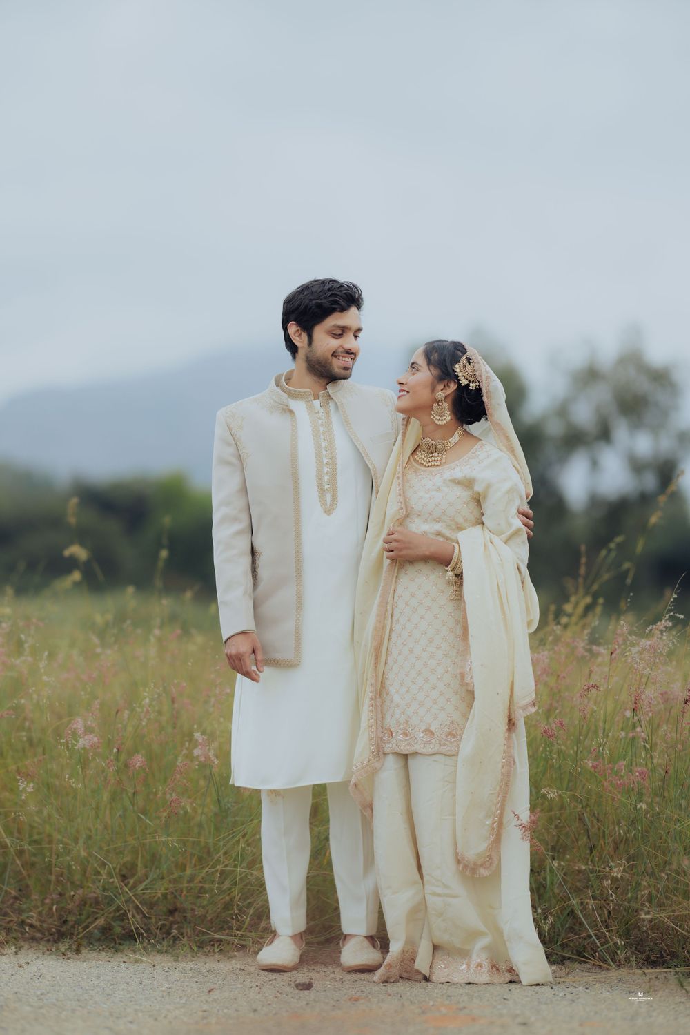 A couple in traditional South Asian wedding attire poses in a field, wearing cream and gold outfits with jewelry.