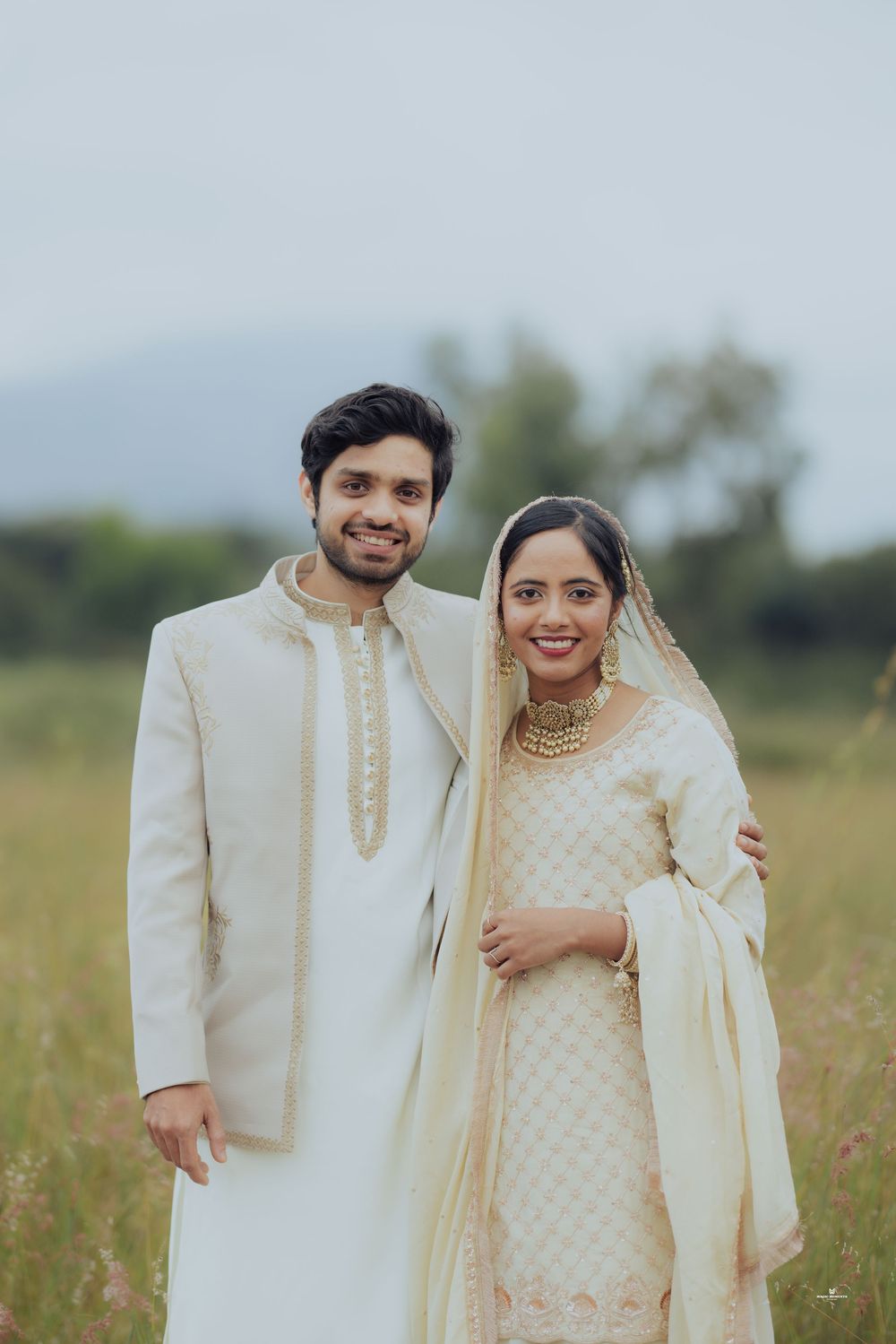 Indian couple in traditional wedding attire posing outdoors, man in white sherwani, woman in cream lehenga with gold jewelry.
