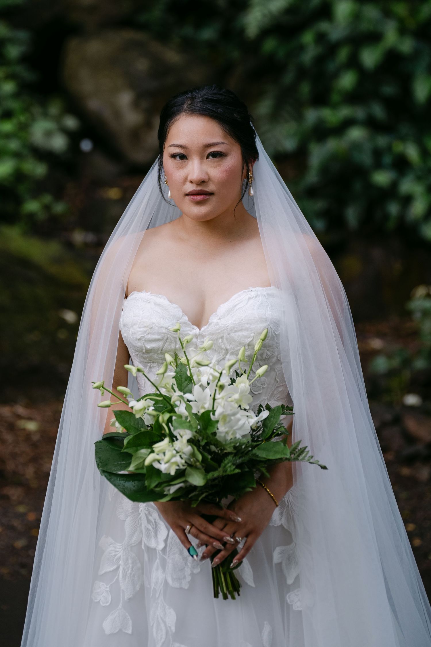 Bride in strapless lace gown and veil holds white floral bouquet outdoors amid lush greenery.