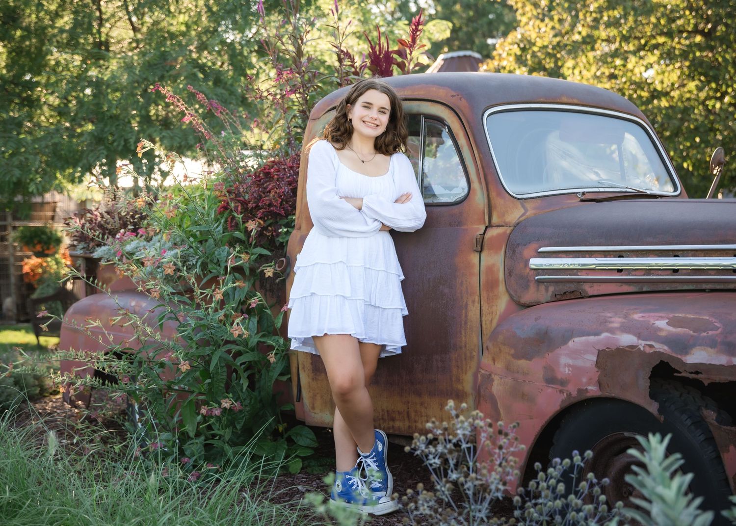 Young woman in white dress and blue sneakers poses beside a rustic vintage truck surrounded by lush garden foliage.