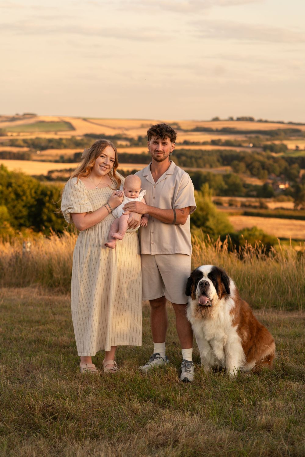 Young family with baby and Saint Bernard dog posing in golden hour sunlight on a scenic hillside meadow.