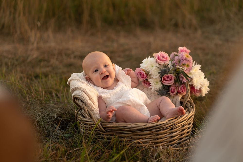 A laughing baby in a white onesie sits in a wicker basket with pink and white flowers in a golden field.