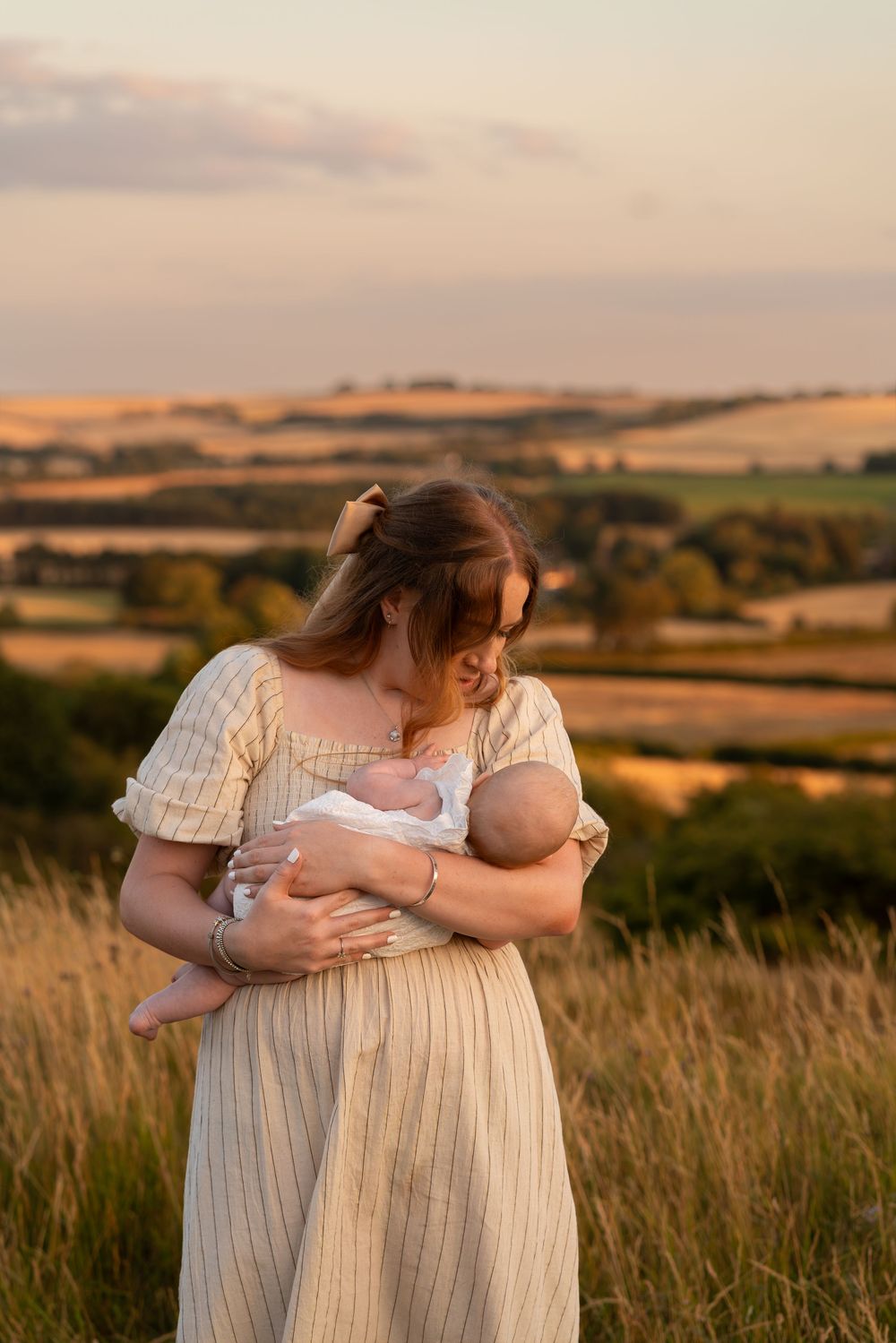 A mother in a striped dress tenderly holds her newborn baby in a golden sunset field with rolling hills.