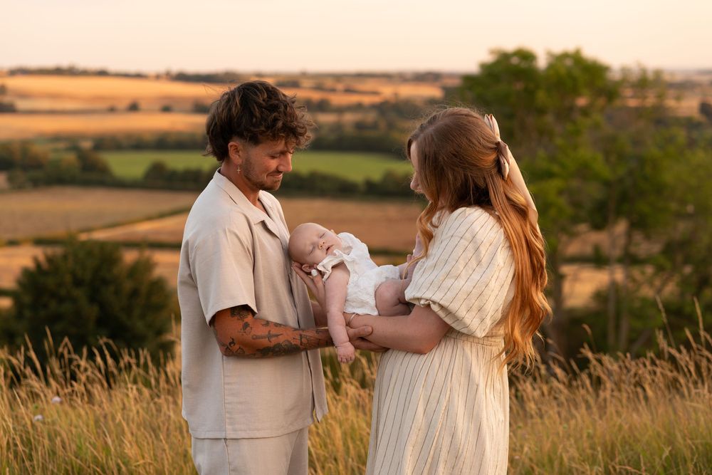 Young couple holding newborn baby in golden field at sunset, warm family portrait outdoors.
