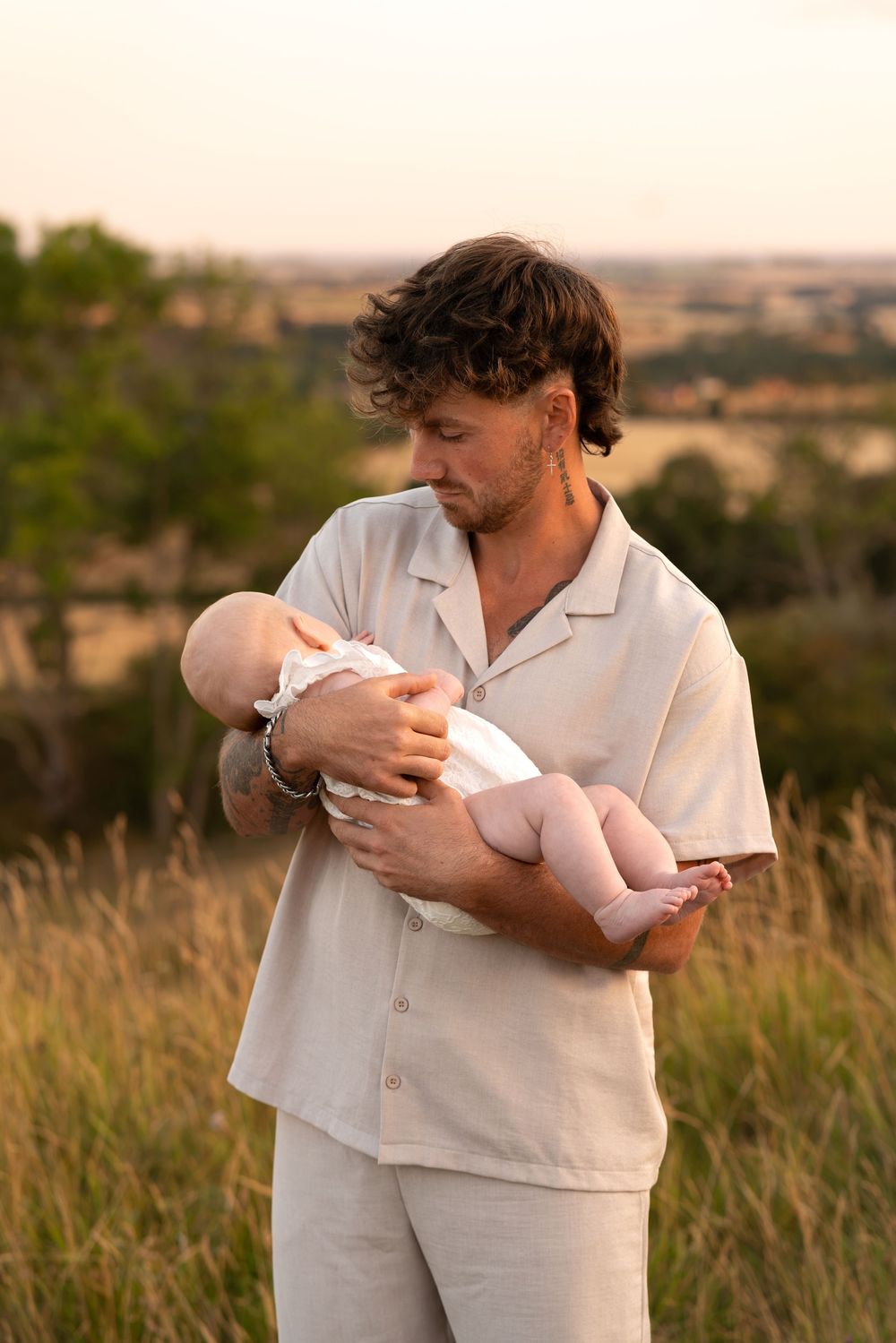 A young man in beige linen outfit tenderly holds a newborn baby in a golden sunset field.