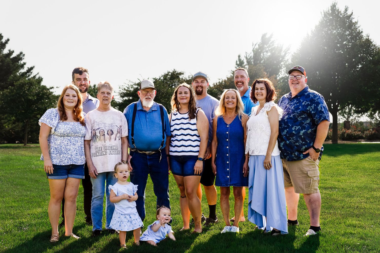 Large family group posing outdoors on green grass in warm sunlight, including adults and a toddler, dressed in casual summer attire.
