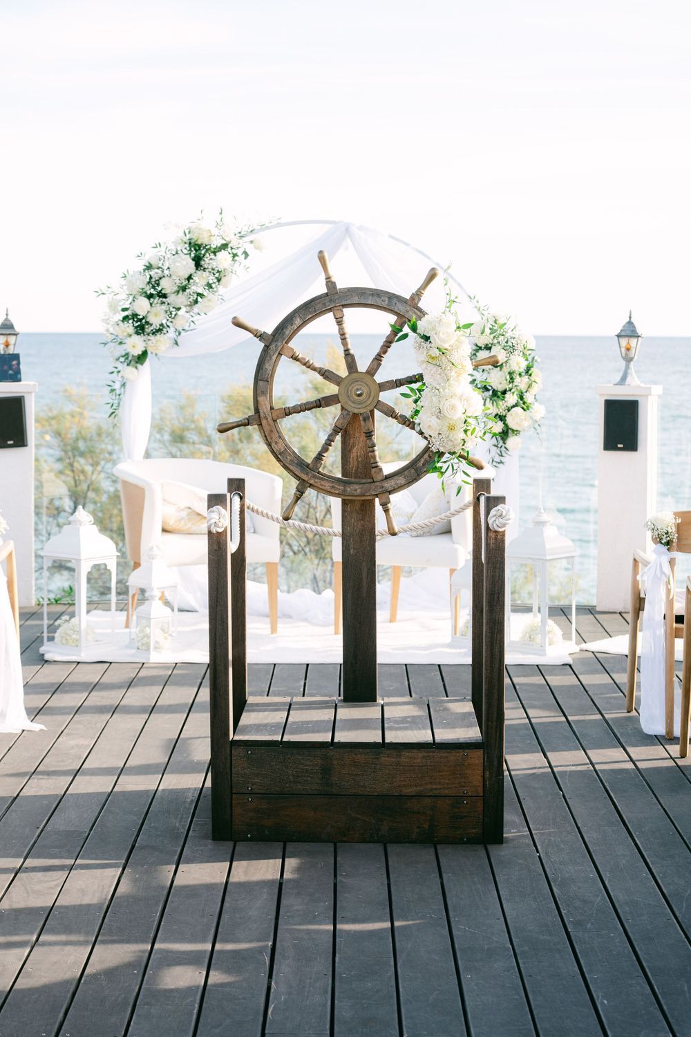 Nautical wedding ceremony decor featuring a wooden ship wheel on a waterfront deck with white floral arch and ocean backdrop.