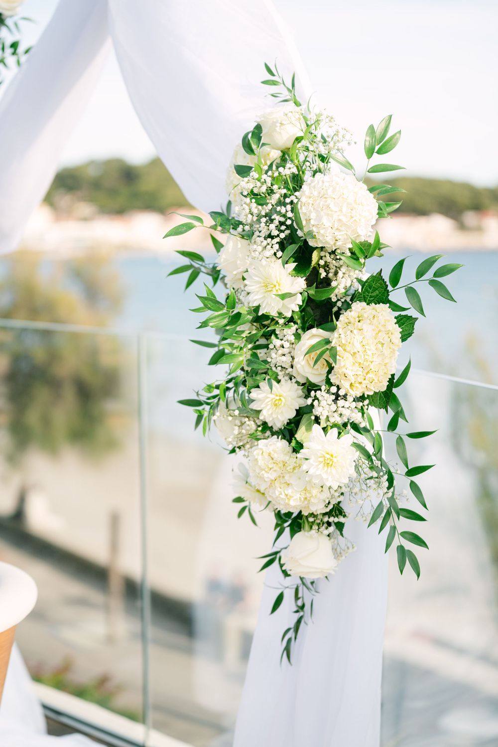 White hydrangea and greenery floral arrangement on draped wedding arch overlooking ocean ceremony venue.
