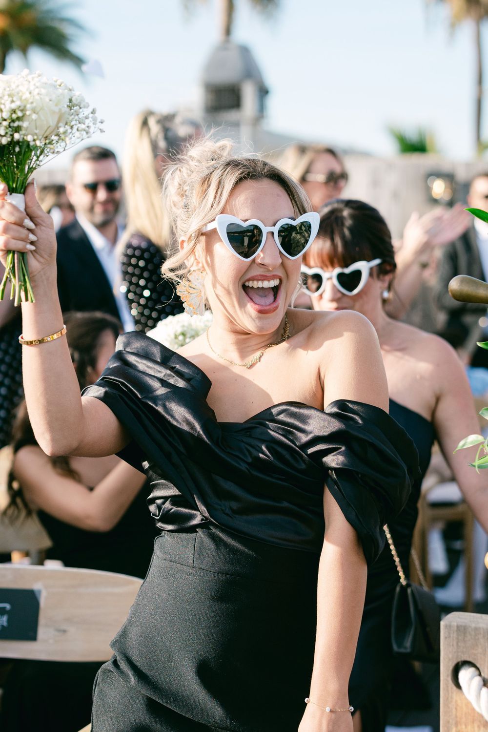 Joyful bridesmaid in black off-shoulder dress and white heart sunglasses holding bouquet at outdoor wedding ceremony.