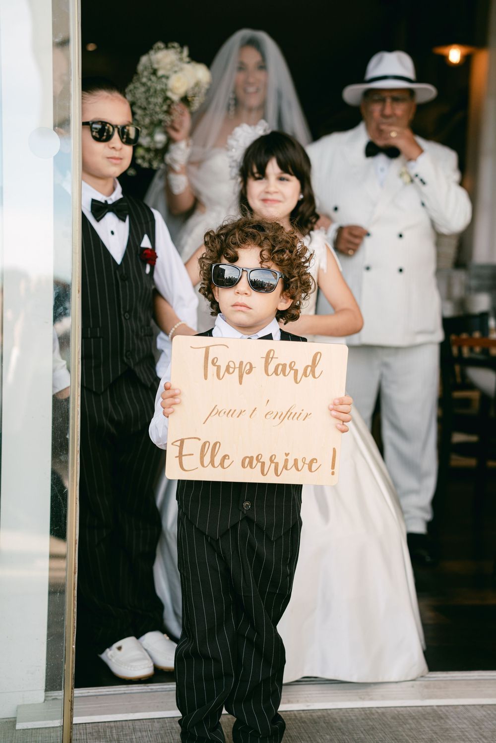 Child ring bearer in pinstripe suit holds French wedding sign, with bride, groom, and children in background.