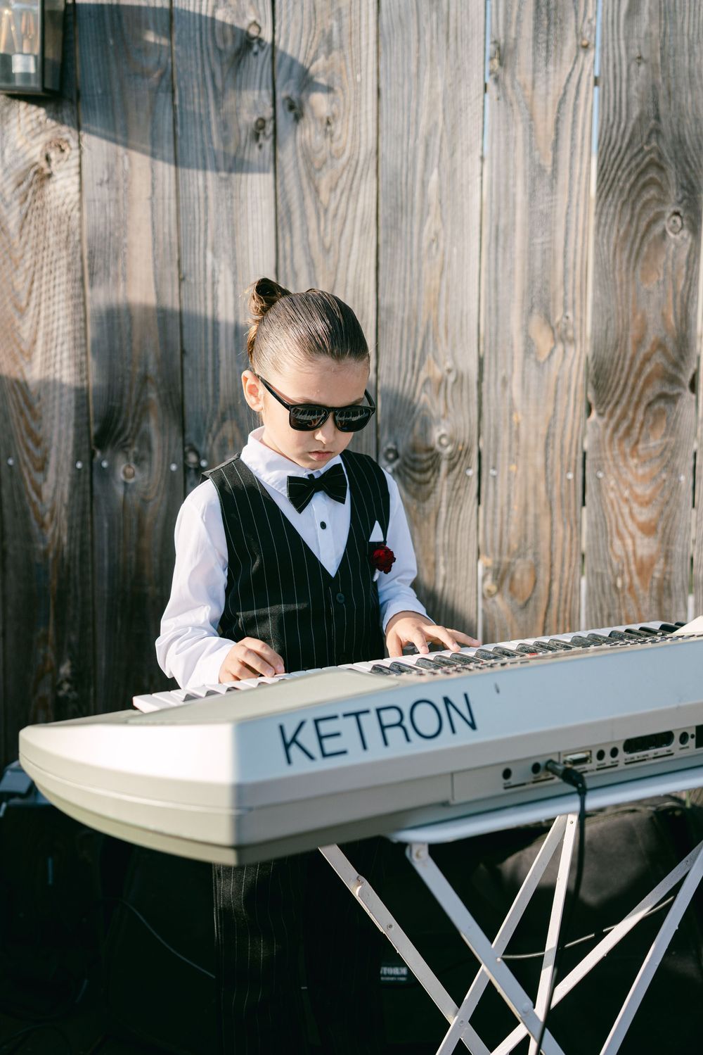 Young boy in tuxedo vest and bow tie playing a Ketron keyboard at an outdoor event.