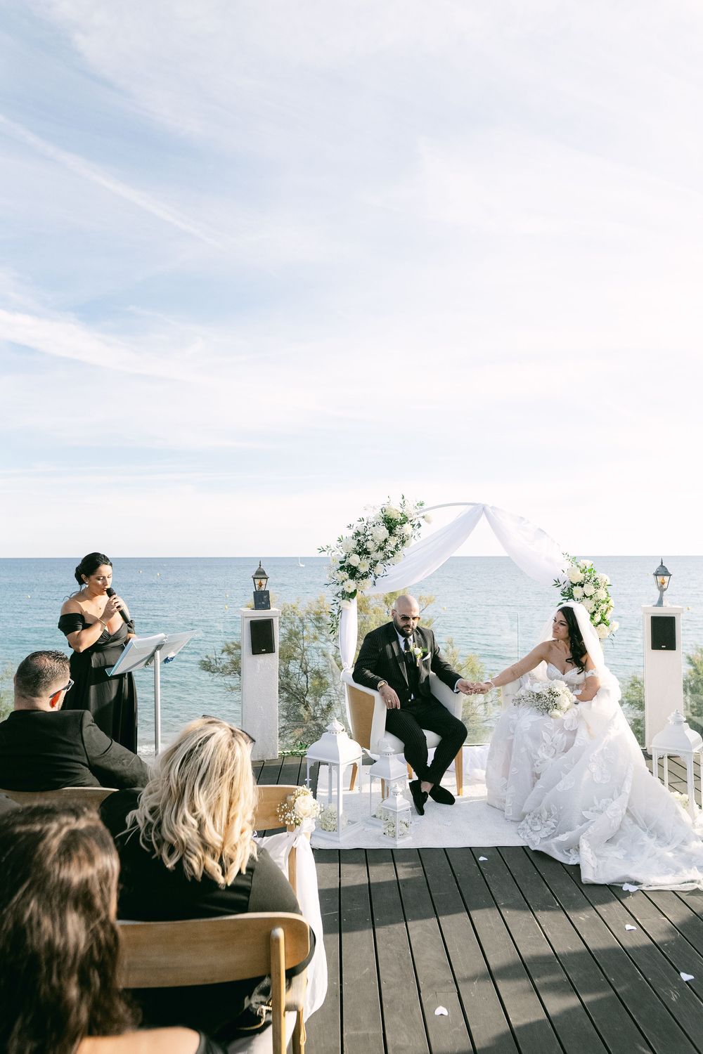 Outdoor wedding ceremony on ocean-view deck with bride and groom seated at floral arch while officiant reads vows.