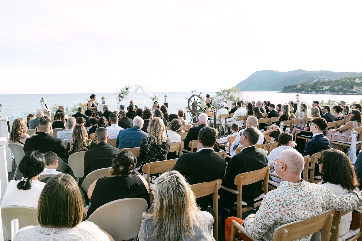 Outdoor wedding ceremony with guests seated in wooden chairs overlooking a scenic ocean and hillside backdrop.